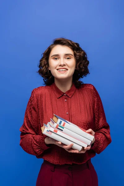 Mujer llena de alegría en blusa roja sosteniendo libros de idiomas y mirando a la cámara aislada en azul - foto de stock