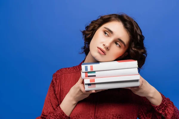Aburrido estudiante sosteniendo libros de texto de idiomas extranjeros mientras mira hacia otro lado aislado en azul - foto de stock