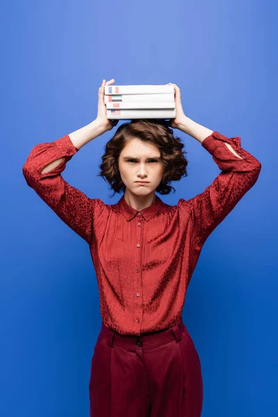 Unzufriedener Student hält fremdsprachige Lehrbücher über dem Kopf und blickt isoliert in die Kamera auf blauem Grund — Stockfoto
