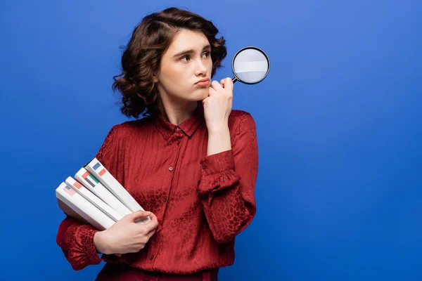 Thoughtful student holding magnifier and textbooks of foreign languages while looking away isolated on blue — Stock Photo
