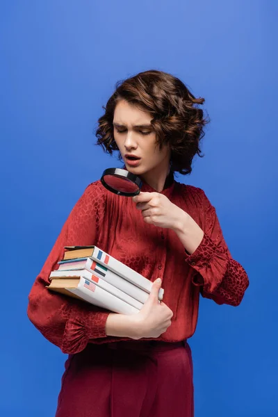 Estudante espantado olhando para dicionários através de lupa isolada em azul — Fotografia de Stock