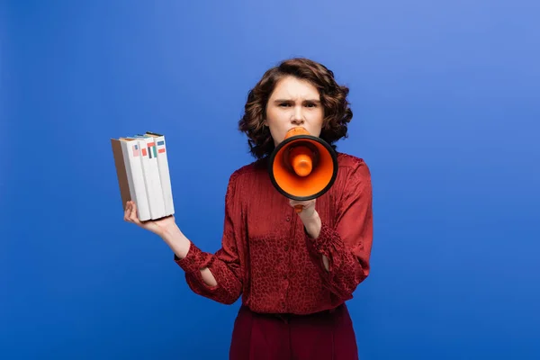 Strict language teacher holding textbooks and screaming in megaphone isolated on blue — Stock Photo