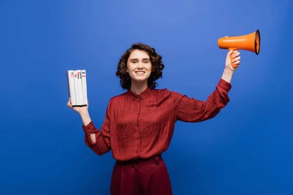 Excited language teacher holding megaphone and textbooks while smiling at camera isolated on blue — Stock Photo