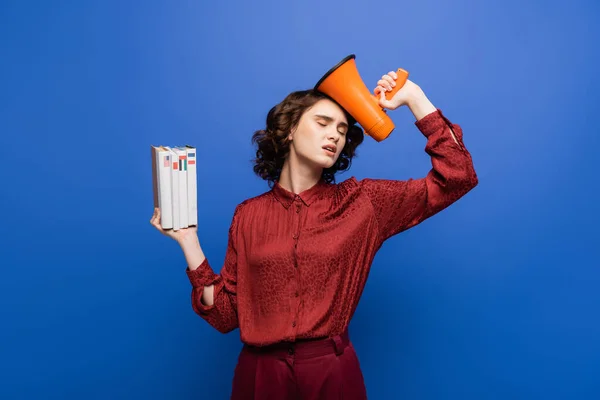 Exhausted language teacher standing with closed eyes while holding megaphone and textbooks isolated on blue — Stock Photo