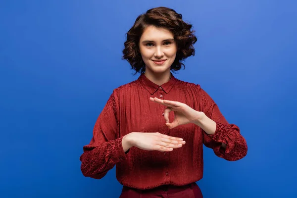 Joyeuse jeune femme souriant à la caméra tout en montrant geste de langue des signes isolé sur bleu — Photo de stock