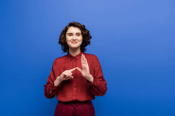 Glückliche junge Lehrerin zeigt Alphabet auf Zeichensprache und blickt vereinzelt in die Kamera auf blauem Grund — Stockfoto