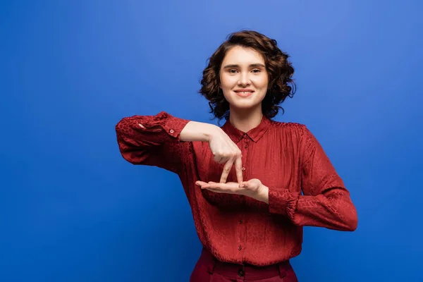 Femme brune souriante et montrant un geste signifiant se tenir debout sur le langage des signes isolé sur bleu — Photo de stock