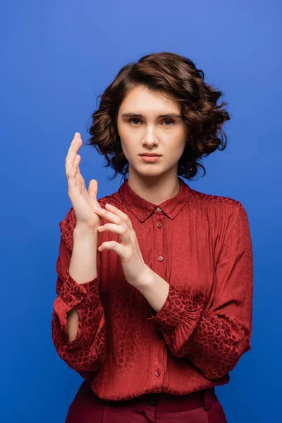 Bonita maestra en blusa roja hablando en lenguaje de señas y mirando a la cámara aislada en azul - foto de stock