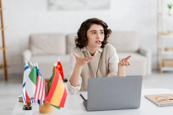 Brunette language teacher in headset gesturing near laptop and blurred international flags at home — Stock Photo
