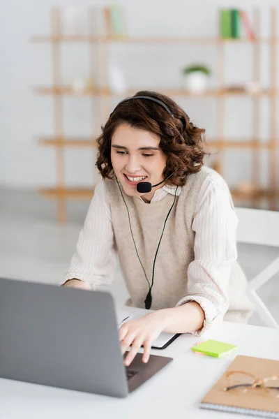 Junge und glückliche Sprachlehrerin tippt im Headset auf Laptop, während sie zu Hause Online-Unterricht hat — Stockfoto