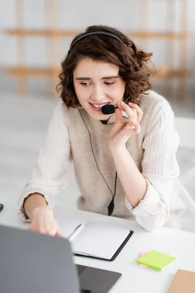 Smiling language teacher in headset talking near laptop and blank notebook while working at home — Stock Photo
