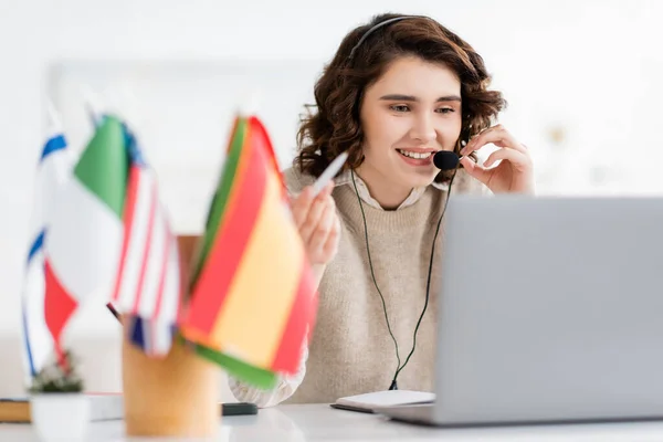 Overjoyed language teacher in headset holding pen and talking near laptop and international flags on blurred foreground — Stock Photo