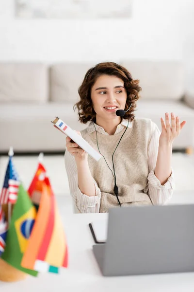 Happy language teacher in headset holding French dictionary while talking during online lesson near international flags — Stock Photo