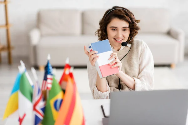Smiling language teacher in headset showing French textbook during video lesson on laptop near blurred international flags — Stock Photo