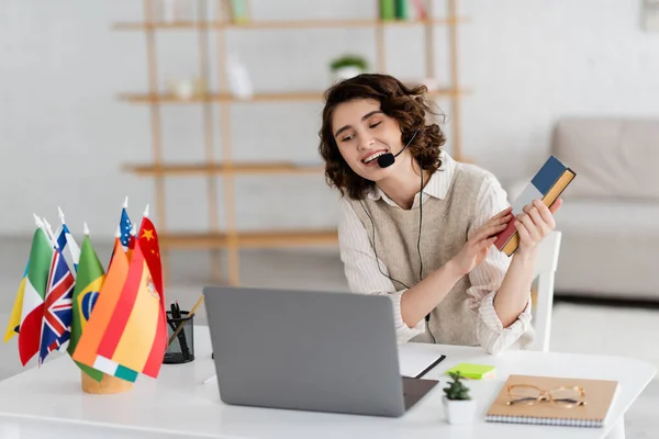 Carefree language teacher in headset holding textbook during online lesson on laptop near international flags — Stock Photo