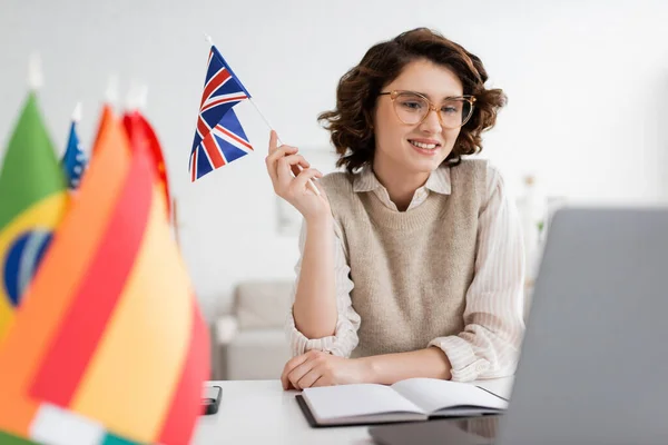 Учитель мови посміхається в окулярах і тримає прапор Сполученого Королівства біля записника і розмитого ноутбука. — стокове фото