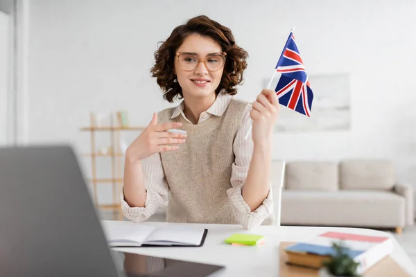 Jeune professeur de langue en lunettes tenant le drapeau du Royaume-Uni près d'un ordinateur portable sur le premier plan flou à la maison — Photo de stock
