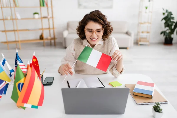 Joyful language teacher showing Italian flag during online lesson on laptop near notebooks and smartphone — Stock Photo
