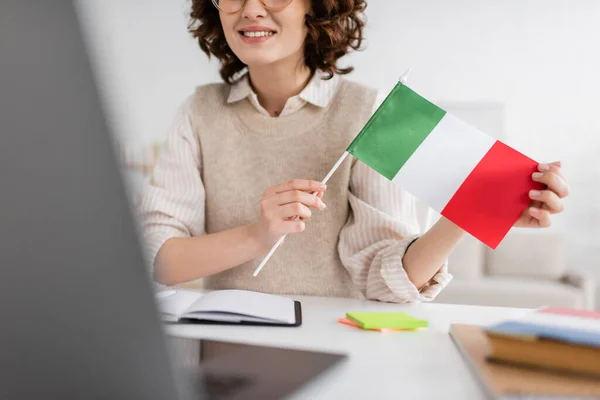 Partial view of smiling language teacher holding flag of Italy during online lesson on blurred laptop at home — Stock Photo