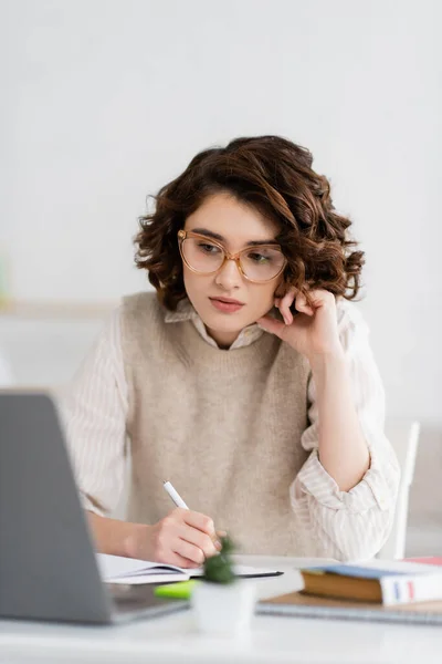 Pretty student in glasses taking notes while learning foreign language and watching online lesson on laptop — Stock Photo