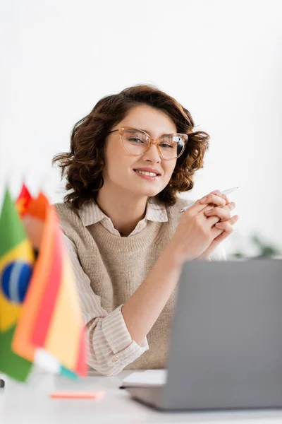 Cheerful language teacher in glasses sitting next to laptop and flags on blurred foreground — Stock Photo