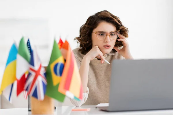 Professor de idiomas concentrado em óculos olhando para laptop perto de bandeiras internacionais em primeiro plano desfocado — Fotografia de Stock