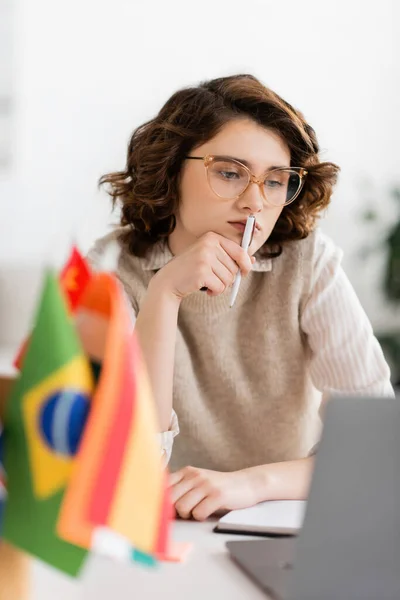 Pensive language teacher in glasses looking at laptop near international flags on blurred foreground — Stock Photo