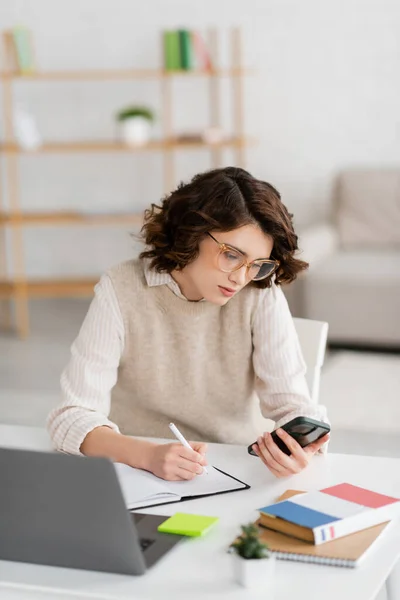 Curly student in glasses holding smartphone while taking notes near laptop at home — Stock Photo