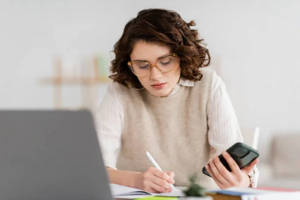 Jolie étudiante en lunettes tenant smartphone tout en prenant des notes près d'un ordinateur portable flou sur le bureau — Photo de stock