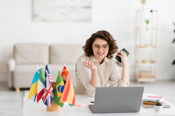 Cheerful language teacher in glasses holding smartphone near laptop and different flags on blurred foreground — Stock Photo