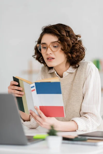 Curly student in glasses reading French dictionary book near laptop on desk — Stock Photo