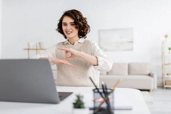 Happy teacher with curly hair showing sign language gesture during online lesson on laptop — Stock Photo