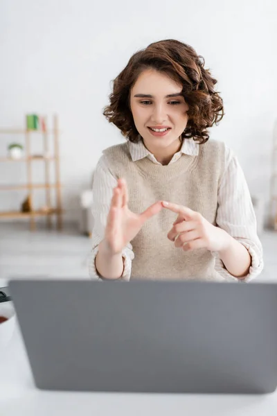 Cheerful woman with curly hair teaching sign language alphabet near blurred laptop at home — Stock Photo