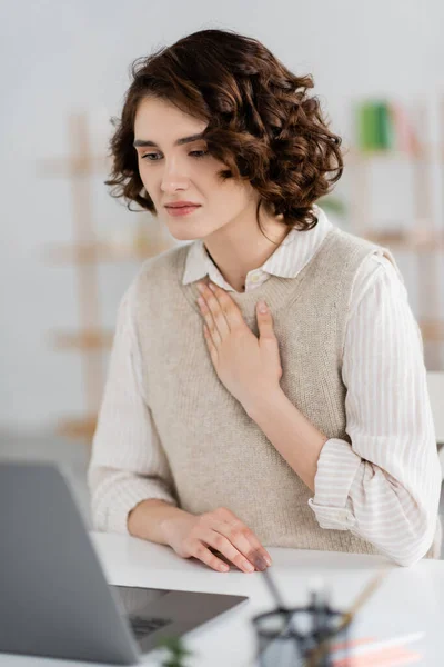 Mujer morena rizada mostrando por favor gesto en el lenguaje de señas mientras que tiene lección en línea en el ordenador portátil borroso en casa - foto de stock