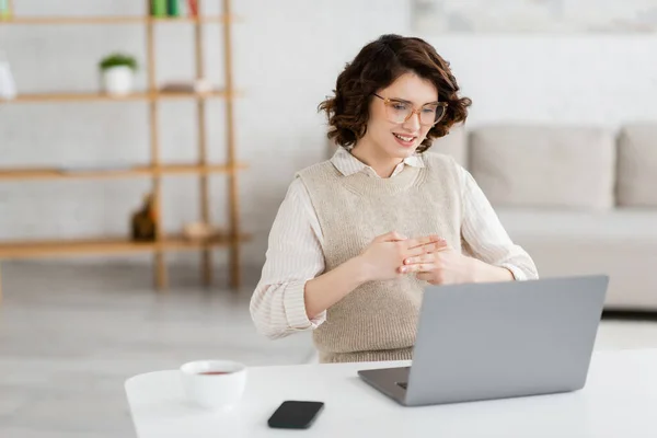 Happy young woman in glasses teaching sign language during online lesson near devices on desk — Stock Photo