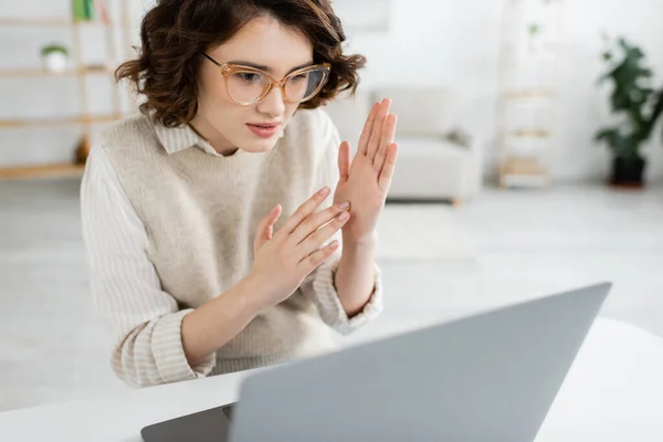 Young teacher in glasses showing two handed sign language gesture while looking at laptop — Stock Photo