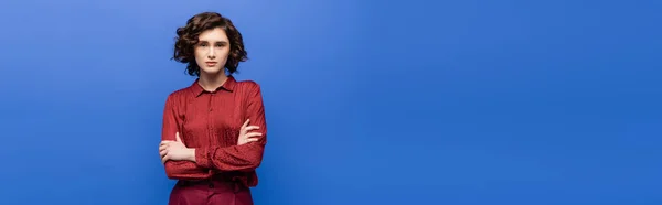 Young language teacher in burgundy blouse standing with crossed arms and looking at camera isolated on blue, banner — Stock Photo
