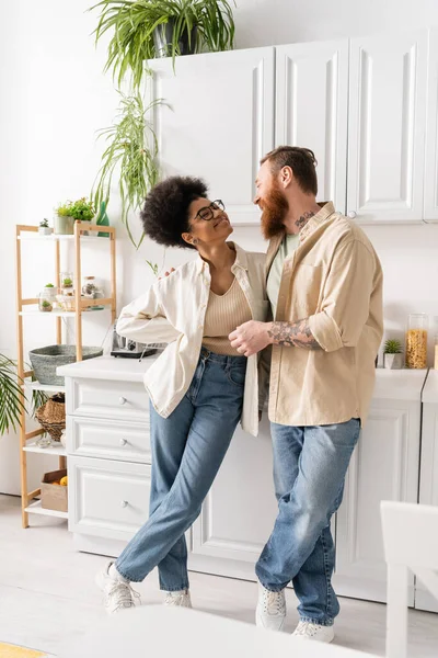 Sonriente pareja interracial mirándose y hablando en la cocina moderna en casa - foto de stock