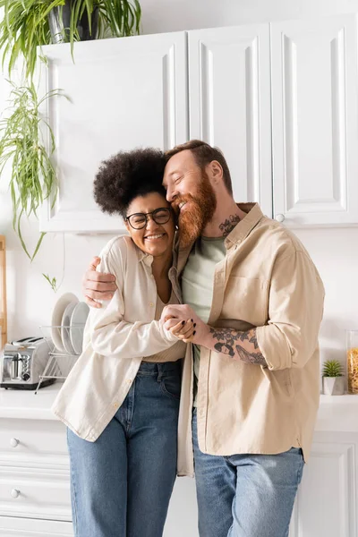 Sonriente hombre tatuado abrazándose y tomándose de la mano con su novia afroamericana en la cocina - foto de stock