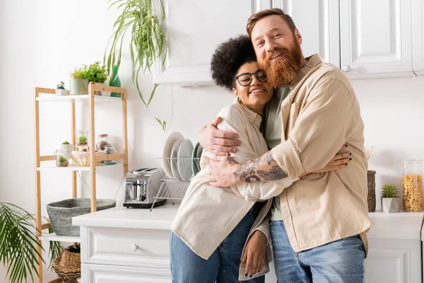 Smiling bearded man hugging african american girlfriend in eyeglasses while standing in kitchen — Stock Photo