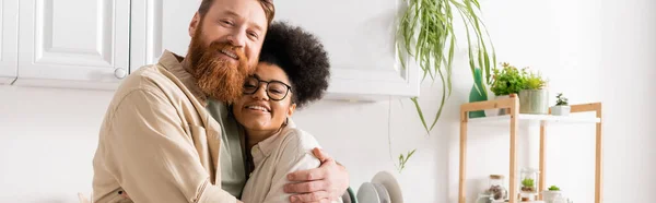 Positive bearded man hugging african american girlfriend in kitchen at home, banner — Stock Photo