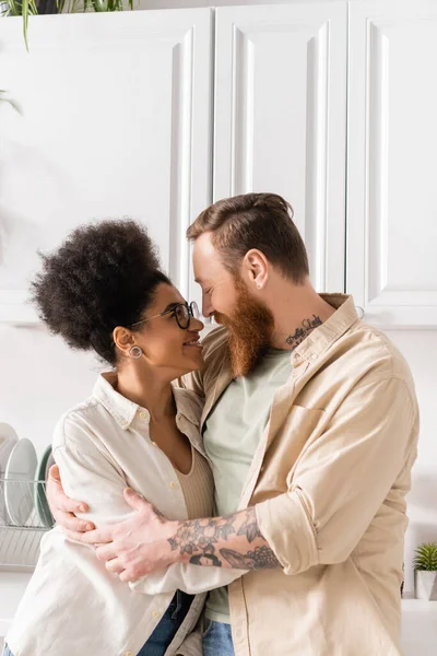 Despreocupada mujer afroamericana abrazando novio tatuado con barba en la cocina - foto de stock