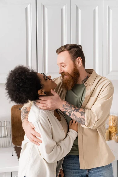 Positif tatoué homme toucher afro-américaine copine dans la cuisine à la maison — Photo de stock