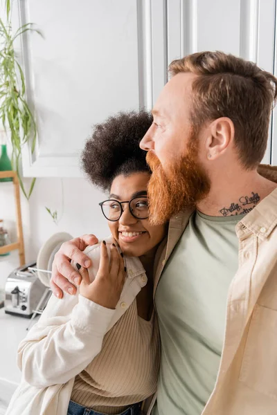 Barbudo homem abraçando alegre afro-americano namorada na cozinha — Fotografia de Stock