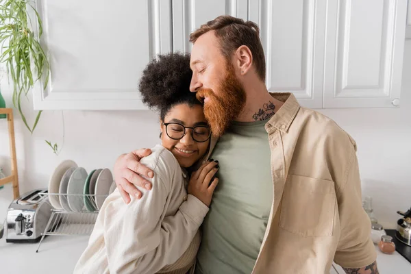 Overjoyed man hugging african american girlfriend in eyeglasses in kitchen — Stock Photo