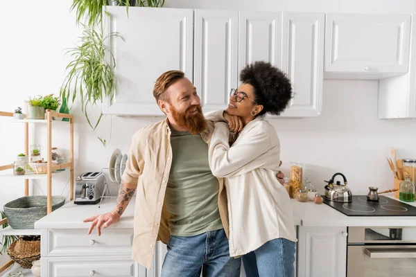 Femme afro-américaine positive regardant petit ami barbu dans la cuisine — Photo de stock