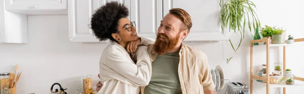 Cheerful african american woman in eyeglasses looking at bearded boyfriend in kitchen, banner — Stock Photo