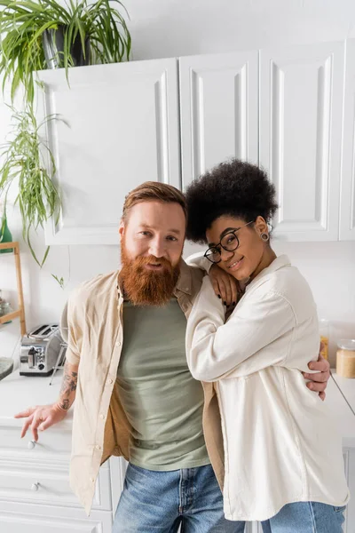 Улыбающийся бородатый мужчина обнимает счастливую африканскую американскую подружку на кухне — стоковое фото