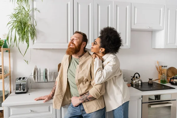 Mujer afroamericana positiva abrazando y hablando con novio barbudo en la cocina - foto de stock
