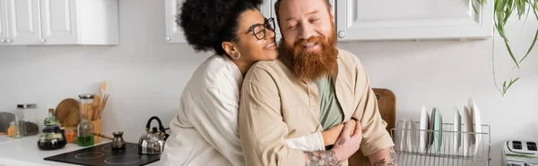 Smiling african american woman hugging tattooed boyfriend in kitchen, banner — Stock Photo